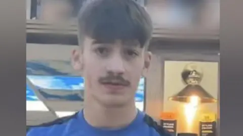 A picture of Taylor wearing a blue shirt with a white Nike logo on it. He has a short moustache and short hair.
