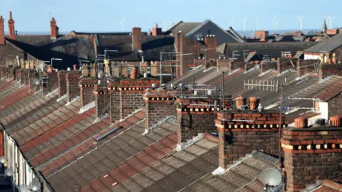 A shot of a row of terraced house roofs, with chimneys, aerials and satellite dishes on display. 