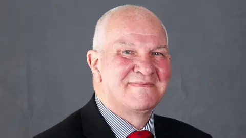 Scottish Borders Council A bald-headed man with short, cropped grey hair smiles into the camera. He is in a dark suit with a striped shirt and a red tie. The background is grey.