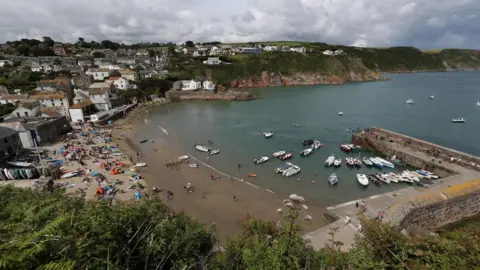 PA Media Gorran Haven harbour viewed from a cliff above it on a cloudy day. The beach is packed with people and watercraft. The calm sea has many swimmers in it and there are about 15 small motorboats moored inside the harbour wall. The village is visible towards the right hand side of the beach and many of the houses are white.
