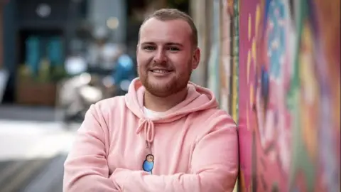 Gary Harper has short light brown hair and a short beard. He is wearing a pink hooded top and is smiling at the camera. He is leaning against a painted wall.