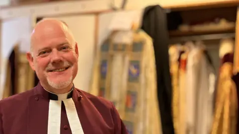 On the left of the photograph, The Very Reverend Joe Hawes, in a red cassock with white clerical collar stands in the foreground in fromt of a wardrobe filled with priest's clothing