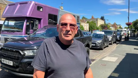 A man with sunglasses standing infront of a high street packed with cars