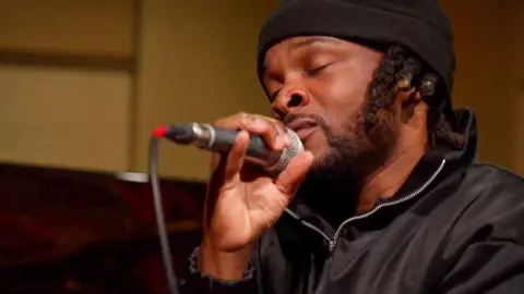 Rosca Onya singing into a microphone. His eyes are closed and he wears a black hat and a black top