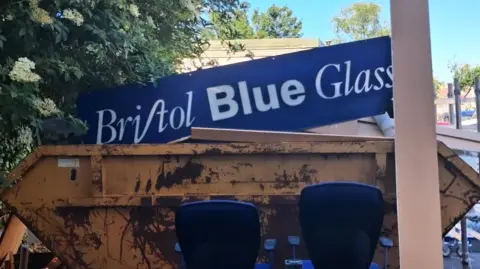 A Bristol Blue Glass shop sign is sat in a skip. The skip is large and has two computer chairs and a plank a wood surrounding it. The sign is partly covered by a bush. 
