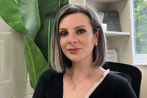 Lucy with a shoulder-length grey bob and a nose ring sitting in her office. It is a head and shoulders shot and there is a green plant behind her.