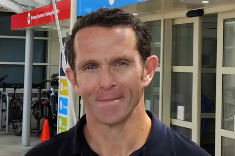 States of Guernsey A man looking into the camera and smiling. He has brown hair and is wearing a navy shirt. He's standing in front of the entrance of a hospital. 