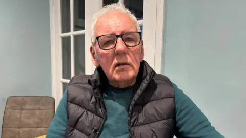 BBC Noel Thomas looks at the camera. He has short white hair and has black-rimmed glasses. He is wearing a blue jumper with a black gilet. A dining table chair can be seen behind him as well as a door and a blue wall. It is a head and shoulders shot.
