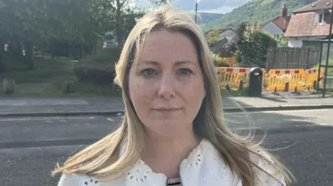 Close-up of a person with light hair wearing a white patterned jacket, standing on a quiet residential street with houses, trees, and green hills in the background under a partly cloudy sky, with orange road barriers and a bin visible behind.