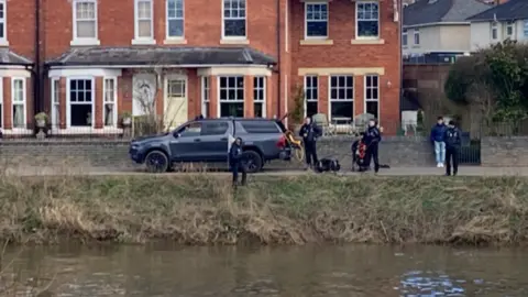 BBC Rescue crews stand by the riverbank in Diglis. Members of the public watch as crews use equipment from a vehicle.