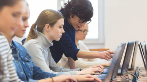 A teacher is leaning over a student who is typing on a laptop. She is wearing glasses and a dark blue shirt and is pointing at her screen.