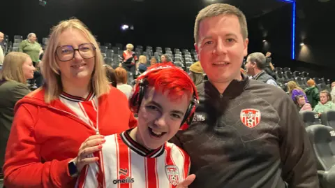 Cian has bright red hair and is seen smiling while wearing red ear protectors.
He is in a Derry City shirt and his parents are standing either side of him.
They are pictured in the movie theatre as a number of people are taking their seats.