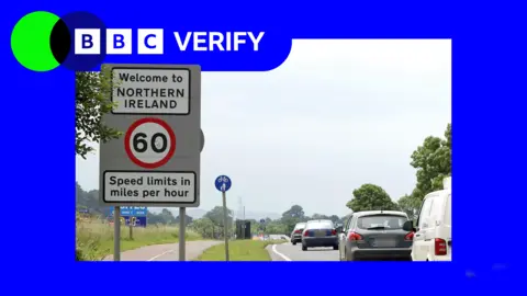 Cars drive towards a sign reading "Welcome to Northern Ireland". 