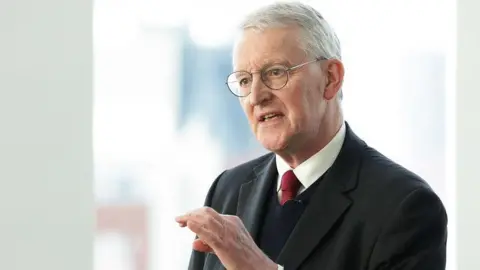 PA Media Hilary Benn has white hair and light framed glasses and is talking with his hand slightly raised. He is wearing a dark suit, dark v-neck jumper, white shirt and red tie.