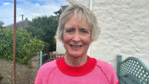 Wendy Kennedy, aged 63 with grey hair, faces the camera smiling. She is wearing a pink pullover with a red neck.