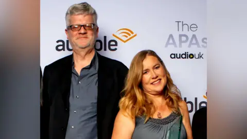 Handout A man in a black suit jacket and grey shirt is standing next to a woman in a dark bottle green dress. They are at an awards ceremony.