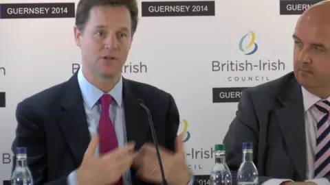 Nick Clegg on the left speaking into a microphone in front of a banner which says British-Irish Council on it. He's wearing a navy blue suit, blue shirt and purple tie. On the right is Deputy Jonathan Le Tocq, a bald man with a grey jacket, white shirt and pink and blue striped tie. 