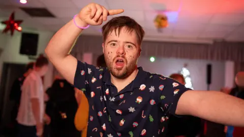 Man is wearing a Christmas themed polo shirt. On his right hand he is wearing a pink glow stick bracelet and has his hand in the air, finger pointing while he's dancing. He has glitter on his cheeks. He has brown, spiky hair. His left arm is out to the left of the image.