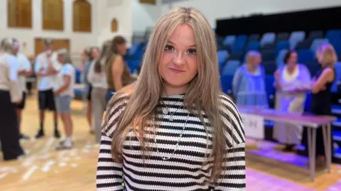 Savanna is wearing a striped black and white top standing in a school hall with others collecting their GCSE results.