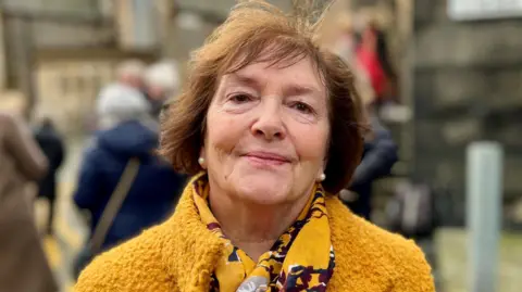 A woman wearing a textured mustard-yellow coat and a patterned yellow scarf with dark and white accents, standing outdoors with blurred buildings and people in the background.