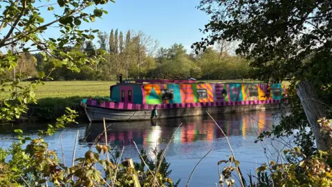 Debbie S A multi-coloured canal boat floating on the water on a sunny day and captured through the trees and bushes.