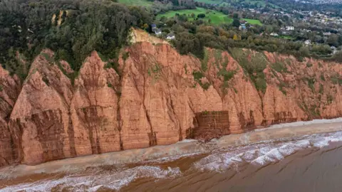 James Loveridge/BNPS A house with a conical roof sits close to the edge above the sea with more houses further inland and nearby hedges and trees along with signs of a cliff fall nearby with mud going into the sea.