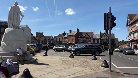 Wantage town Market Place car park is in the centre of town with a road that runs around it. In this photo it is very busy with a full car park. A stone statue of King Alfred stands in the middle of the car park.