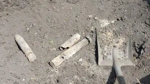 South Wales Police Three unexploded bombs among soil and rubble, they are white in colour and appear corroded. A shovel can be seen on the right hand side of them. 