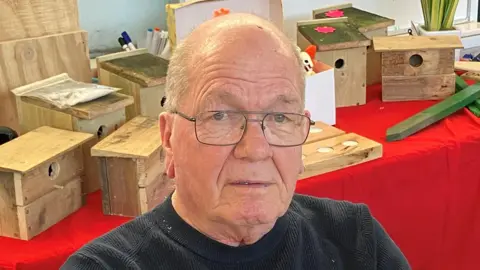 An elderly man, with short grey hair and rectangular glasses, is looking at the camera. He is wearing a black jumper.