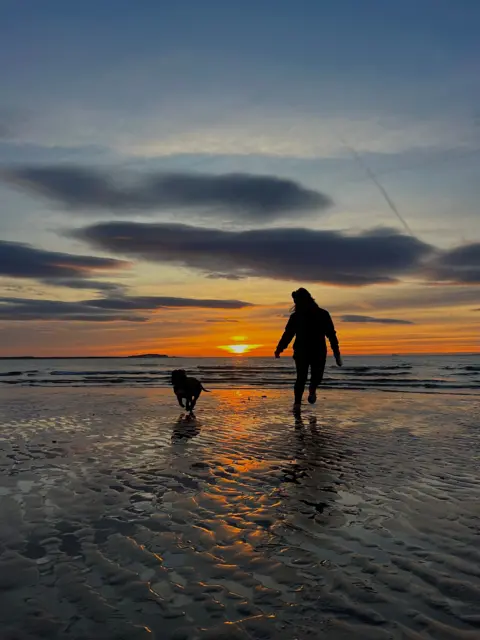 Laura Docherty Sunset reflections across wet sand as a silhouetted women and dog run along the shore.