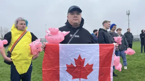 Connor Bennett/BBC A man wearing a blue hoody with the hood up and a blue baseball cap. He is holding up a red and white Canadian flag with a red maple leaf. He is holding an inflatable pig. Behind him are other men holding inflatable pigs standing on grass under grey skies at Witham Town FC. 