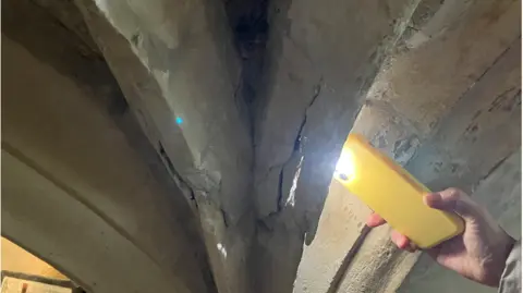 A vaulted ceiling in a medieval crypt is cracked. The light from a mobile phone lights up the gaps in the vault showing it is in poor condition. 