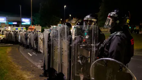 A line of police in riot gear. Their faces are covered. 
