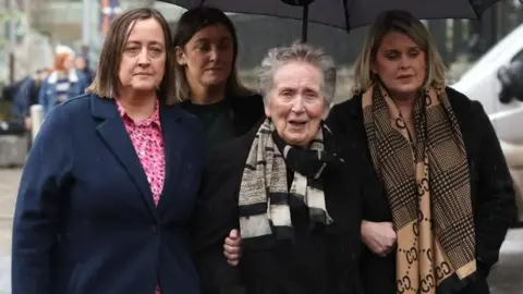 PA Media Four members of the family are standing underneath a large black umbrella. They have sad facial expressions and are all wearing coats and scarves.  