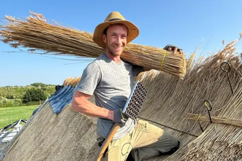 Harrison Rodwell Luke is kneeling on a thatched roof, with long straw thatch tied together placed over his left shoulder. He is wearing a sun hat, grey t-shirt and tan-coloured combat trousers. There are blue skies above.