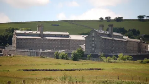 A picture taken from the outside of Dartmoor prison. There is a large grass field to the front of the image. The building is all brick with a number of windows.
