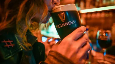 A close-up, side-on view of a woman sipping a pint of guinness. She has shoulder-length blonde hair and is holding the pint in her right hand, holding it to her mouth as she takes the first sip.