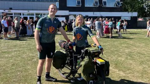 Lucy and Ciaran stand together with a bike that is loaded with panniers, wearing matching, green cycling jerseys. It is a sunny day and they are standing on grass, with a crowd of people mingling in the background.