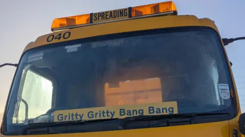 BCP Council Front cab of yellow gritting vehicle with "Gritty, Gritty, Bang, Bang" displayed in its windscreen.