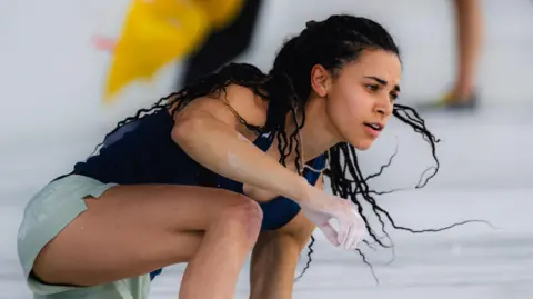 International Federation of Climbing/ Lena Drapella Molly Thompson-Smith. Molly is a 26-year-old woman with long braided hair. She wears a dark blue vest and green shorts and crouches on a crash mat, her right hand covered in chalk, after completing a climb