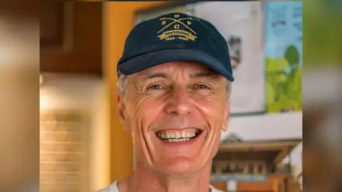 A man with white hair smiling at the camera. He is wearing a navy-coloured hat.