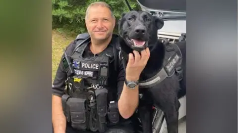 A policeman sits with a dog outside. The man has short grey hair and is wearing a black police uniform, including a vest with pockets for items such as walkie-talkies. The dog has short black fur. The man is tickling the dog under the chin. 