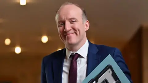 Getty Images a smiling man with a receding fair hair is wearing a blue suit jacket, white shirt and purple tie