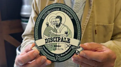 A close up of an ale label which would be seen on a beer tap, being held by a man's fingers. On the picture is a drawing of two religious figures with cloaks and halos, each holding a beer and embracing each other. The label reads "DISCIPALE", intended as a pun on 'Disciple' and 'Ale'. It also says 'bitter' and has an ABV of 3.7 per cent. 