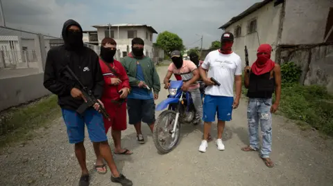 Lee Durant/BBC Seven young men in balaclavas brandish automatic weaponry in broad daylight, facing the camera. two of the men are on a dirt bike. 