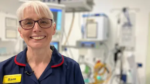 Sam Backway is smiling at the camera. She has white hair, which is tied back so you can only see her fringe. She is wearing a navy nurses uniform which has red piping around the colour and a yellow name tag over her right chest. She has dark framed glasses on. 