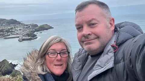 Helen Moorefield David Griffiths and Helen Moorefield dressed in outdoor clothing and standing by the sea. Both are smiling. 