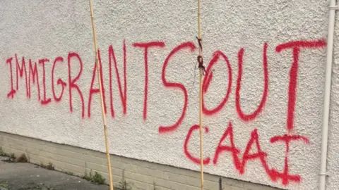 Shows a gable wall with the words Immigrants Out CAAI written in red paint