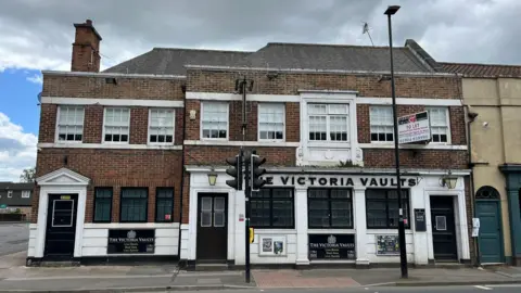 A large brick building is shown across a road. There are traffic lights and a streetlight in the foreground, and the lower half of the building is painted white. The sign says 'The Victoria Vaults' and has large windows. There's three doors to the building, and it has 14 windows in total. There are aerials on the roof and a chimney, as well as a dark green door to the right of the photo which is part of a connecting building. 