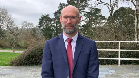 Laurie wears a dark blue suit with red tie and white shirt and stands outside the entrance to Lys Kernow with trees and bushes in the background.